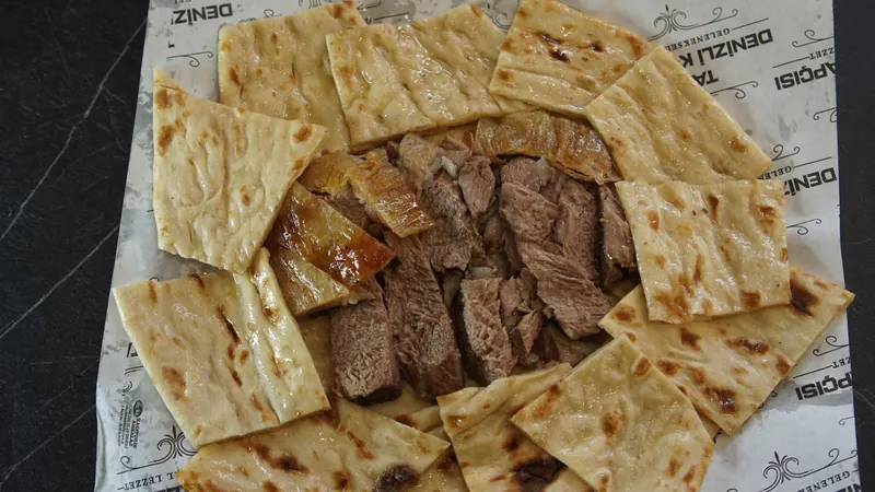 Tarihi Denizli Kebapçısı logolu kâğıt üzerinde pidelerle çevrelenmiş Denizli kebabı, üstten görünüm